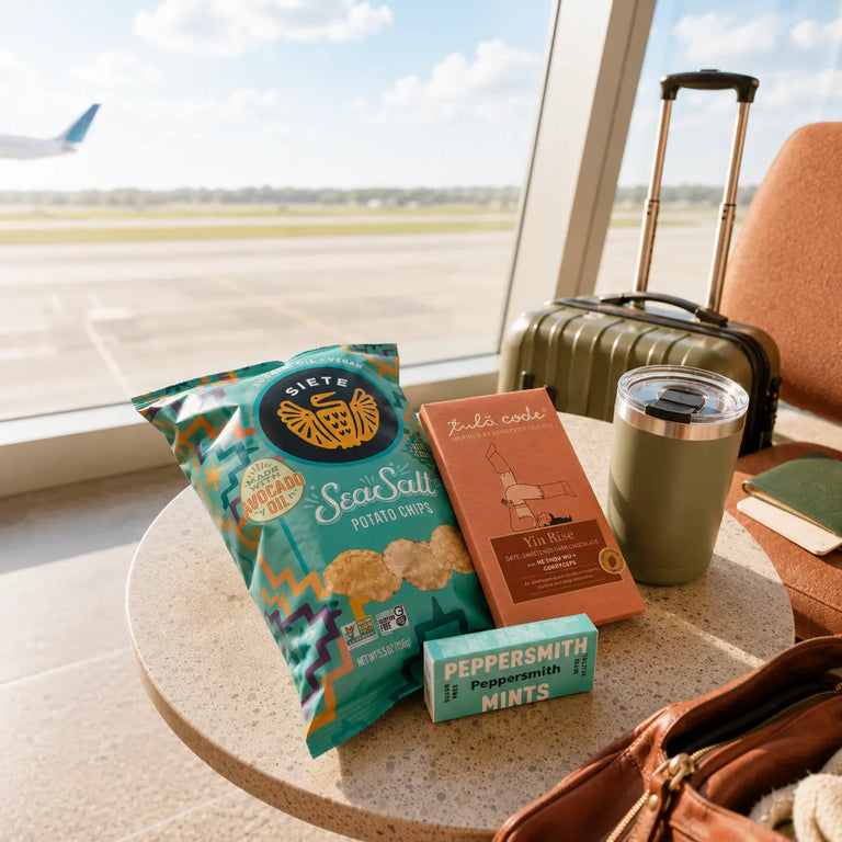 A travel snack setup on a round table by a window, featuring a bag of Piete Sea Salt Potato Chips, a box of Zaku code Tea Time, a green tumbler, and a green Peppermint Mints tin, with a rolling suitcase and an airport tarmac visible in the background.