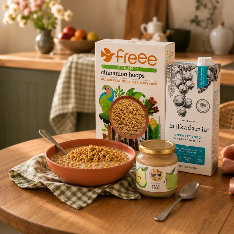 A styled breakfast scene on a wooden table with a bowl of cereal, a spoon, a jar of peanut butter, a carton of milk, and a box of Free Organic cinnamon hoops. The background shows a softly blurred kitchen interior with flowers and fruit.